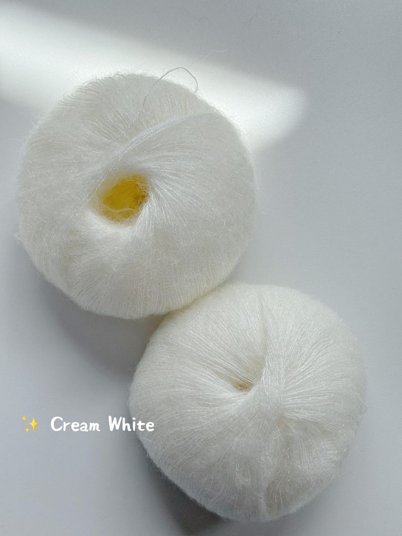 Puede incluir: Dos ovillos de hilo blanco crema se muestran sobre un fondo blanco. El hilo parece suave y esponjoso, con un ligero brillo. El texto "Cream White" es visible en la imagen, indicando el color del hilo.