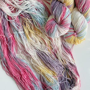Puede incluir: Primer plano de ovillos de lana y hebras sueltas en tonos rosa, morado, amarillo, blanco y azul. La lana est&aacute; dispuesta sobre una superficie blanca, mostrando las vibrantes variaciones de color y texturas, ideal para proyectos de tejido o ganchillo.