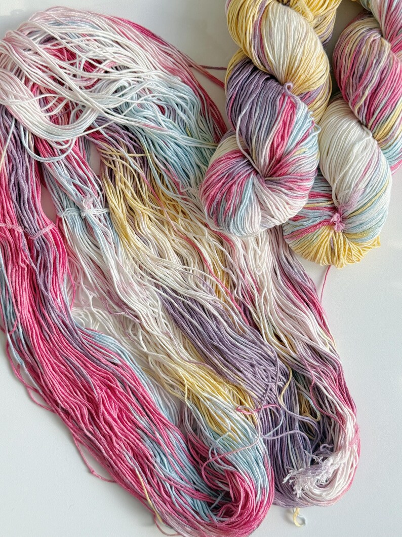 Puede incluir: Primer plano de ovillos de lana y hebras sueltas en tonos rosa, morado, amarillo, blanco y azul. La lana est&aacute; dispuesta sobre una superficie blanca, mostrando las vibrantes variaciones de color y texturas, ideal para proyectos de tejido o ganchillo.