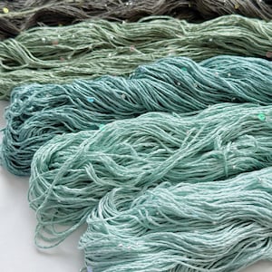 May include: Four skeins of yarn arranged in a gradient from dark charcoal to light mint green. Each strand features small, reflective sequins embedded throughout the texture. The text "Option A" is printed in the bottom left corner of the image.