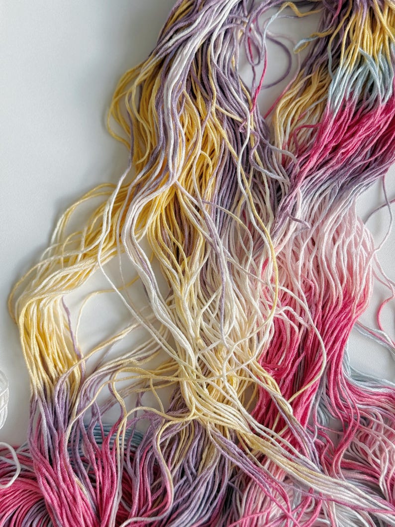 Puede incluir: Primer plano de hilo de bordar colorido en tonos amarillos, rosas, morados y blancos. Los hilos est&aacute;n dispuestos en un haz suelto y enredado, creando una exhibici&oacute;n texturizada y vibrante. Ideal para proyectos de manualidades y bordado.