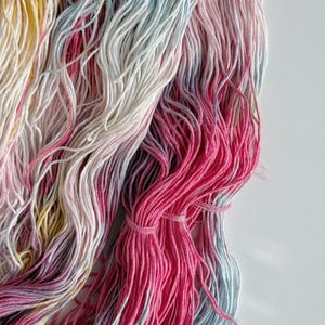 Puede incluir: Primer plano de madejas de hilo coloridas. El hilo es una mezcla de rosa, amarillo, azul y blanco. El hilo est&aacute; agrupado con una peque&ntilde;a banda. El hilo es suave y parece estar hecho de una fibra natural.