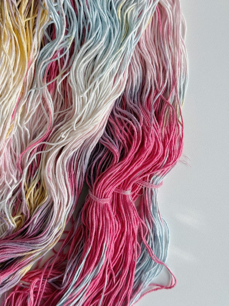 Puede incluir: Primer plano de madejas de hilo coloridas. El hilo es una mezcla de rosa, amarillo, azul y blanco. El hilo est&aacute; agrupado con una peque&ntilde;a banda. El hilo es suave y parece estar hecho de una fibra natural.