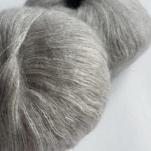 Puede incluir: Primer plano de dos madejas de hilo Moonlit Silver. El hilo es de un color gris claro y suave con un brillo sutil. La textura del hilo es visible, con fibras individuales que crean una apariencia esponjosa. El texto "Moonlit Silver" se muestra en la parte inferior derecha.