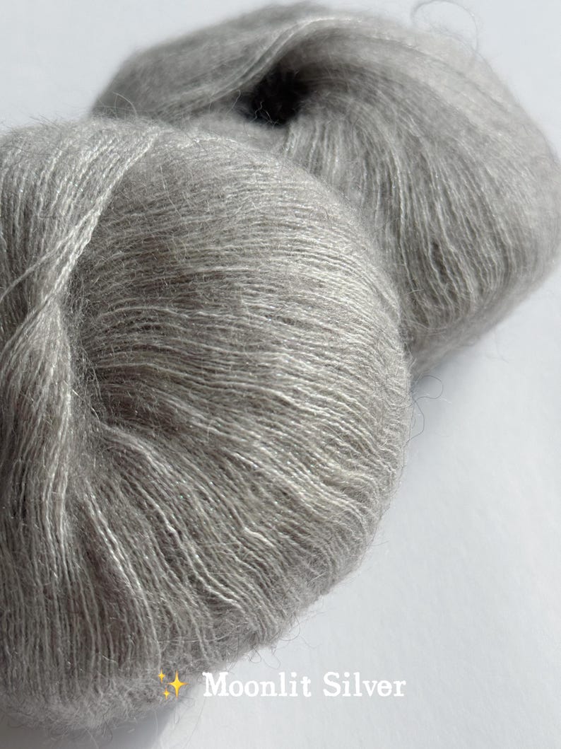 Puede incluir: Primer plano de dos madejas de hilo Moonlit Silver. El hilo es de un color gris claro y suave con un brillo sutil. La textura del hilo es visible, con fibras individuales que crean una apariencia esponjosa. El texto "Moonlit Silver" se muestra en la parte inferior derecha.