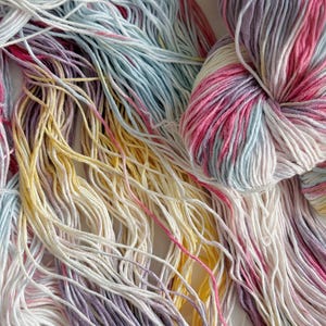 Puede incluir: Primer plano de madejas de hilo coloridas. El hilo es una mezcla de blanco, rosa, amarillo, azul claro y morado. El hilo es suave y esponjoso, perfecto para proyectos de tejido o ganchillo. Los colores se mezclan, creando un efecto hermoso y &uacute;nico.