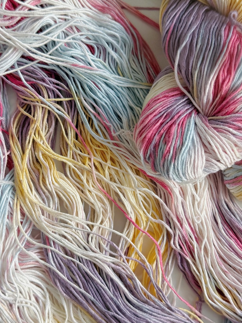 Puede incluir: Primer plano de madejas de hilo coloridas. El hilo es una mezcla de blanco, rosa, amarillo, azul claro y morado. El hilo es suave y esponjoso, perfecto para proyectos de tejido o ganchillo. Los colores se mezclan, creando un efecto hermoso y &uacute;nico.