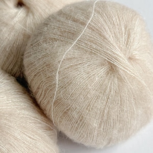 Puede incluir: Primer plano de varias madejas de hilo beige. El hilo tiene una textura suave y esponjosa y un ligero brillo. El texto "Champagne Glow" es visible en la esquina inferior derecha.