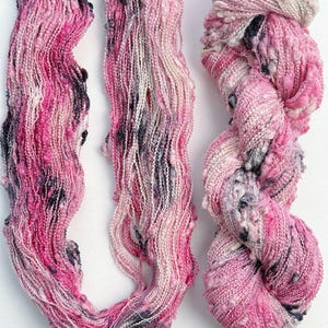 May include: Two skeins of textured art yarn featuring a blend of pink, white, and dark charcoal gray fibers. The yarn has a varied, bumpy, and irregular construction with thick and thin sections throughout the twisted and loose strands.