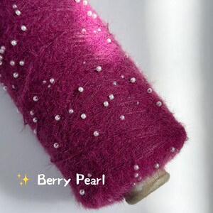 May include: Close-up of a spool of berry-colored yarn adorned with small, white pearls. The yarn has a fuzzy texture, and the pearls are scattered across the surface. The text "Berry Pearl" is visible on the image.