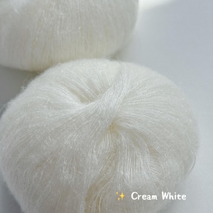 Puede incluir: Dos ovillos de lana color crema. La lana es suave y parece estar hecha de una fibra delicada. El texto "Cream White" se muestra en la parte inferior de la imagen.