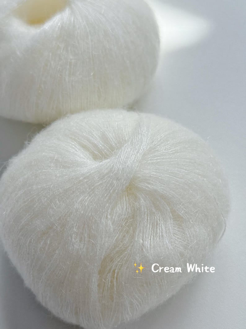 Puede incluir: Dos ovillos de lana color crema. La lana es suave y parece estar hecha de una fibra delicada. El texto "Cream White" se muestra en la parte inferior de la imagen.