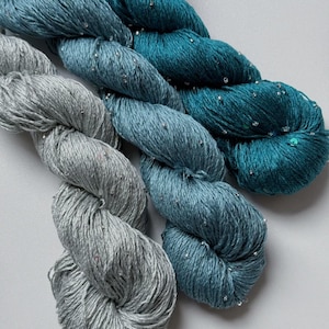 May include: Close-up of several skeins of yarn in shades of teal, blue, and gray. The yarn has a subtle sheen and is speckled with tiny, sparkling embellishments. The arrangement suggests a craft or textile project.