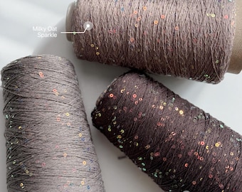 May include: Three spools of yarn featuring a thin thread with small, reflective sequins. The colors are labeled as Milky Oat Sparkle, Espresso Sparkle, and Muted Taupe. Each spool is cylindrical with a cardboard core, showcasing a textured finish.
