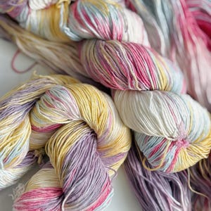 Puede incluir: Primer plano de madejas de hilo coloridas en tonos de amarillo, rosa, morado y azul. El hilo est&aacute; torcido y dispuesto en un patr&oacute;n visualmente atractivo, lo que sugiere un proyecto de artesan&iacute;a o textil. Los colores pastel suaves crean una est&eacute;tica suave y acogedora.