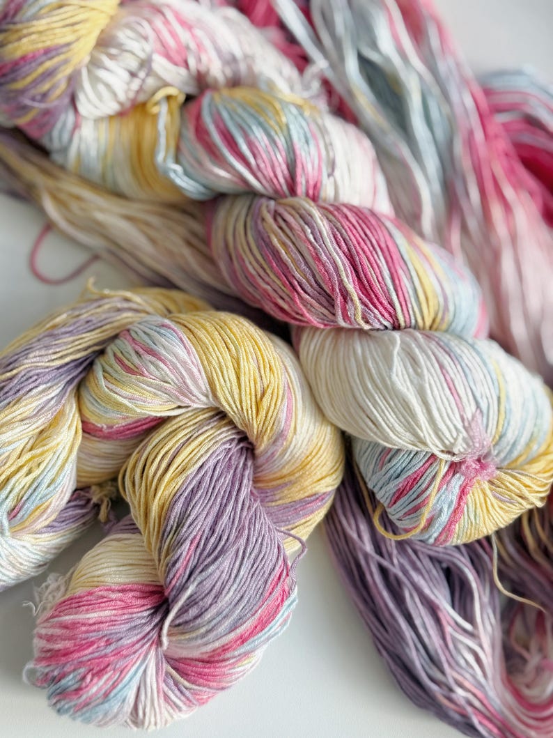 Puede incluir: Primer plano de madejas de hilo coloridas en tonos de amarillo, rosa, morado y azul. El hilo est&aacute; torcido y dispuesto en un patr&oacute;n visualmente atractivo, lo que sugiere un proyecto de artesan&iacute;a o textil. Los colores pastel suaves crean una est&eacute;tica suave y acogedora.