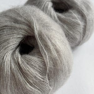 Puede incluir: Dos madejas de hilo Moonlit Silver. El hilo es de un color gris claro y suave con un brillo sutil. El texto "Moonlit Silver" se muestra en blanco en la esquina inferior derecha de la imagen. El hilo parece ser de un peso fino, adecuado para proyectos delicados.