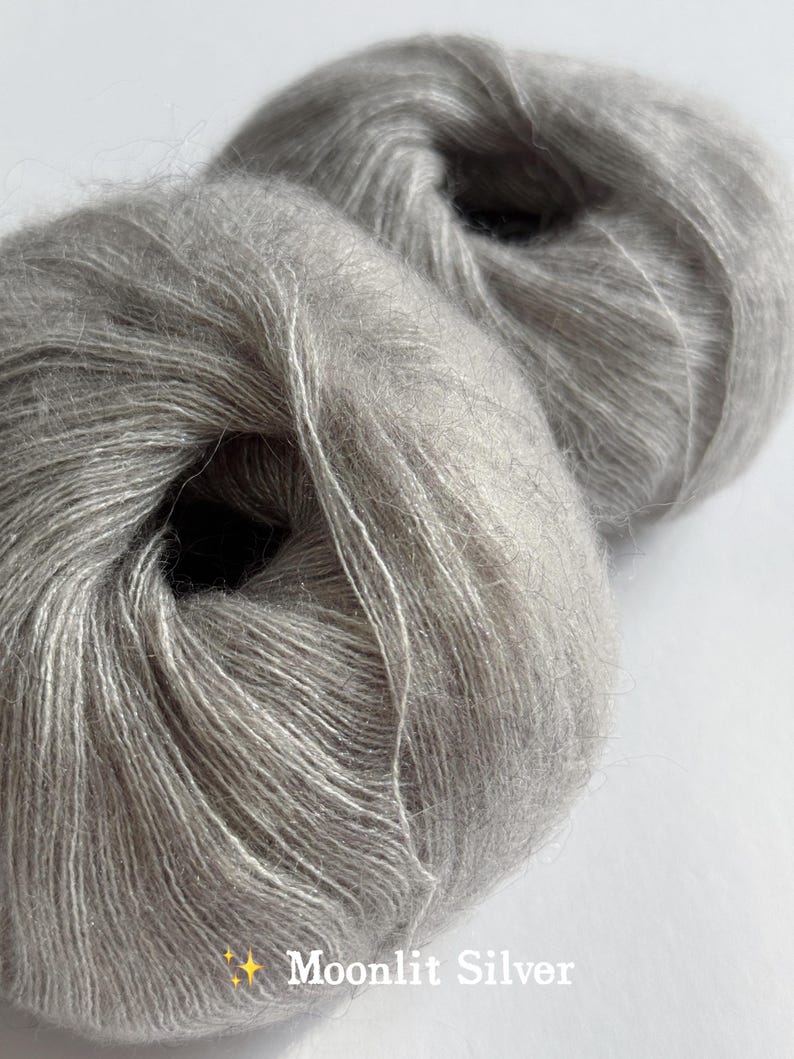 Puede incluir: Dos madejas de hilo Moonlit Silver. El hilo es de un color gris claro y suave con un brillo sutil. El texto "Moonlit Silver" se muestra en blanco en la esquina inferior derecha de la imagen. El hilo parece ser de un peso fino, adecuado para proyectos delicados.