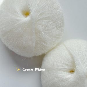 Puede incluir: Dos ovillos de lana blanco crema se muestran sobre un fondo blanco. La lana parece suave y esponjosa, con hebras visibles. El texto "Cream White" est&aacute; presente en la imagen, indicando el color de la lana. Ideal para proyectos de punto y ganchillo.