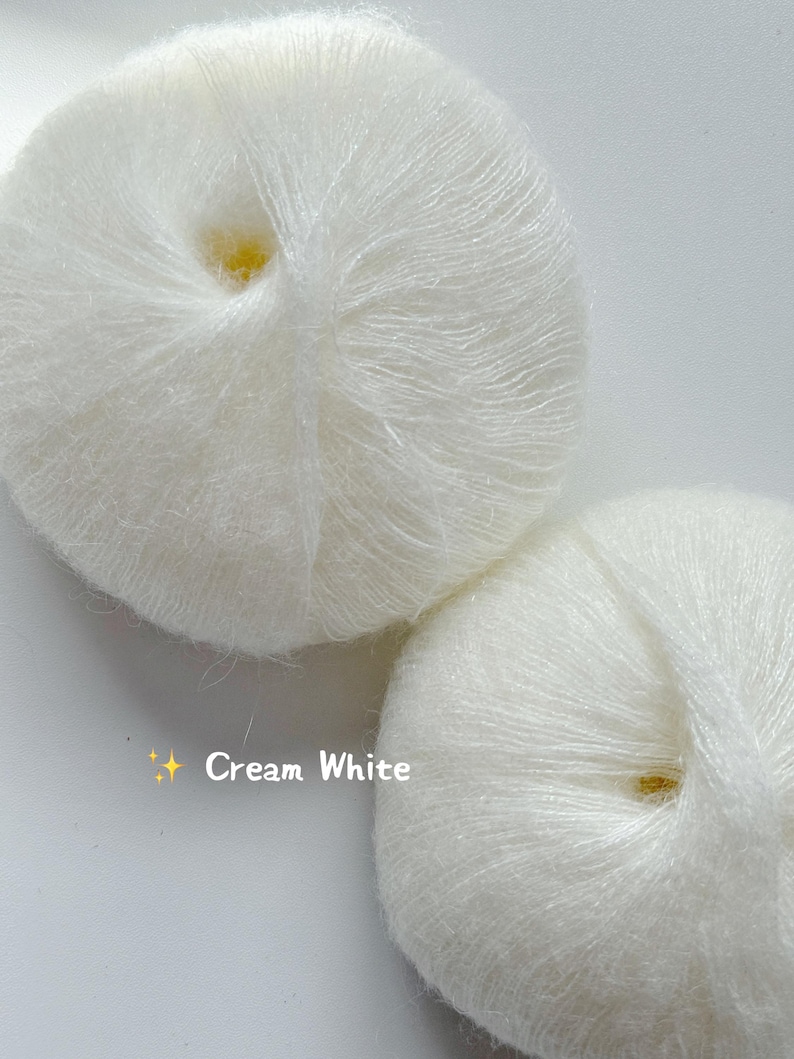 Puede incluir: Dos ovillos de lana blanco crema se muestran sobre un fondo blanco. La lana parece suave y esponjosa, con hebras visibles. El texto "Cream White" est&aacute; presente en la imagen, indicando el color de la lana. Ideal para proyectos de punto y ganchillo.