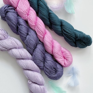 Peut inclure: Vue aérienne de plusieurs écheveaux de fil aux tons rose, violet et sarcelle. Le fil est torsadé et disposé en diagonale. Des plumes aux couleurs pastel sont dispersées autour du fil, ajoutant une touche délicate. Le fond est blanc uni.