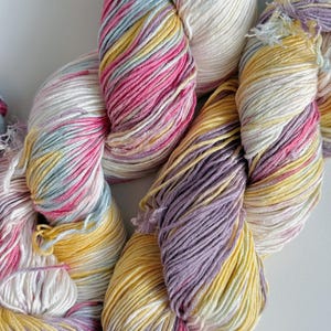 Puede incluir: Primer plano de madejas de hilo coloridas. El hilo es una mezcla de blanco, rosa, amarillo y lavanda. El hilo es suave y parece estar hecho de una fibra natural. Las madejas est&aacute;n dispuestas en un patr&oacute;n suelto y fluido.