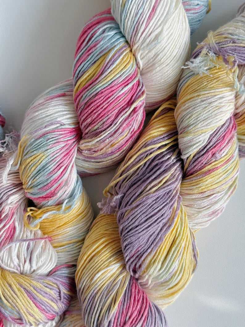 Puede incluir: Primer plano de madejas de hilo coloridas. El hilo es una mezcla de blanco, rosa, amarillo y lavanda. El hilo es suave y parece estar hecho de una fibra natural. Las madejas est&aacute;n dispuestas en un patr&oacute;n suelto y fluido.