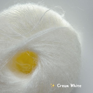 Puede incluir: Primer plano de una madeja de hilo blanco crema. El hilo es esponjoso y suave, con una textura delicada. El texto "Cream White" se muestra en la esquina inferior derecha.