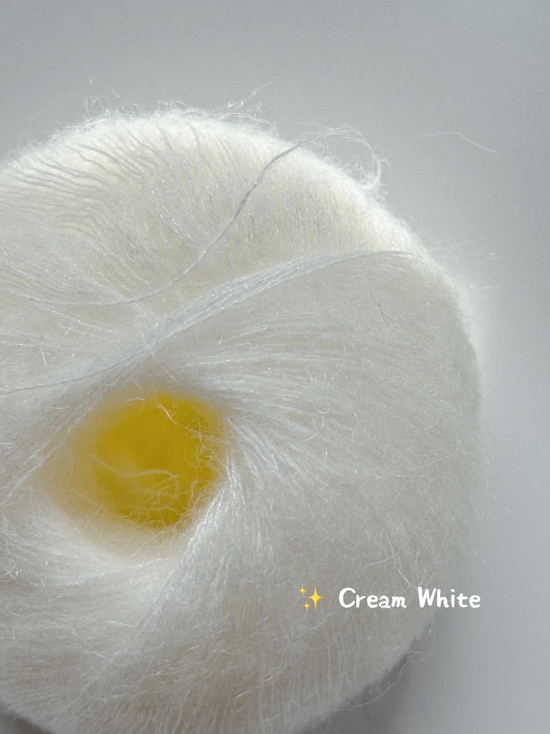 Puede incluir: Primer plano de una madeja de hilo blanco crema. El hilo es esponjoso y suave, con una textura delicada. El texto "Cream White" se muestra en la esquina inferior derecha.