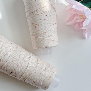May include: Two spools of off-white macrame cord with flecks of red, yellow, and orange. The cord is wrapped around a clear plastic base. A pink flower and green leaves are in the background.