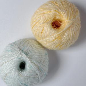 May include: Two balls of yarn. One is a pale yellow with hints of orange, and the other is a light blue with flecks of teal. The yarn is soft and fluffy, ideal for knitting or crocheting projects.