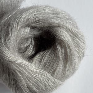 Puede incluir: Primer plano de un ovillo de hilo Moonlit Silver. El hilo es de un color gris claro y suave con un brillo sutil. El texto "Moonlit Silver" se muestra en la parte inferior de la imagen. Ideal para proyectos de punto y ganchillo.