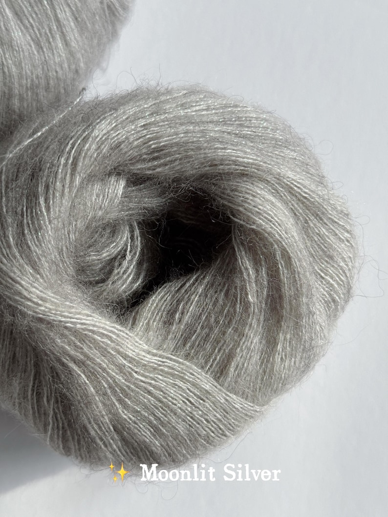 Puede incluir: Primer plano de un ovillo de hilo Moonlit Silver. El hilo es de un color gris claro y suave con un brillo sutil. El texto "Moonlit Silver" se muestra en la parte inferior de la imagen. Ideal para proyectos de punto y ganchillo.