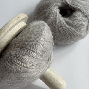 Puede incluir: Dos madejas de hilo Moonlit Silver, una en un carrete de madera. El hilo es de un color gris claro y suave, con una textura esponjosa. El texto "Moonlit Silver" est&aacute; en la parte inferior de la imagen.