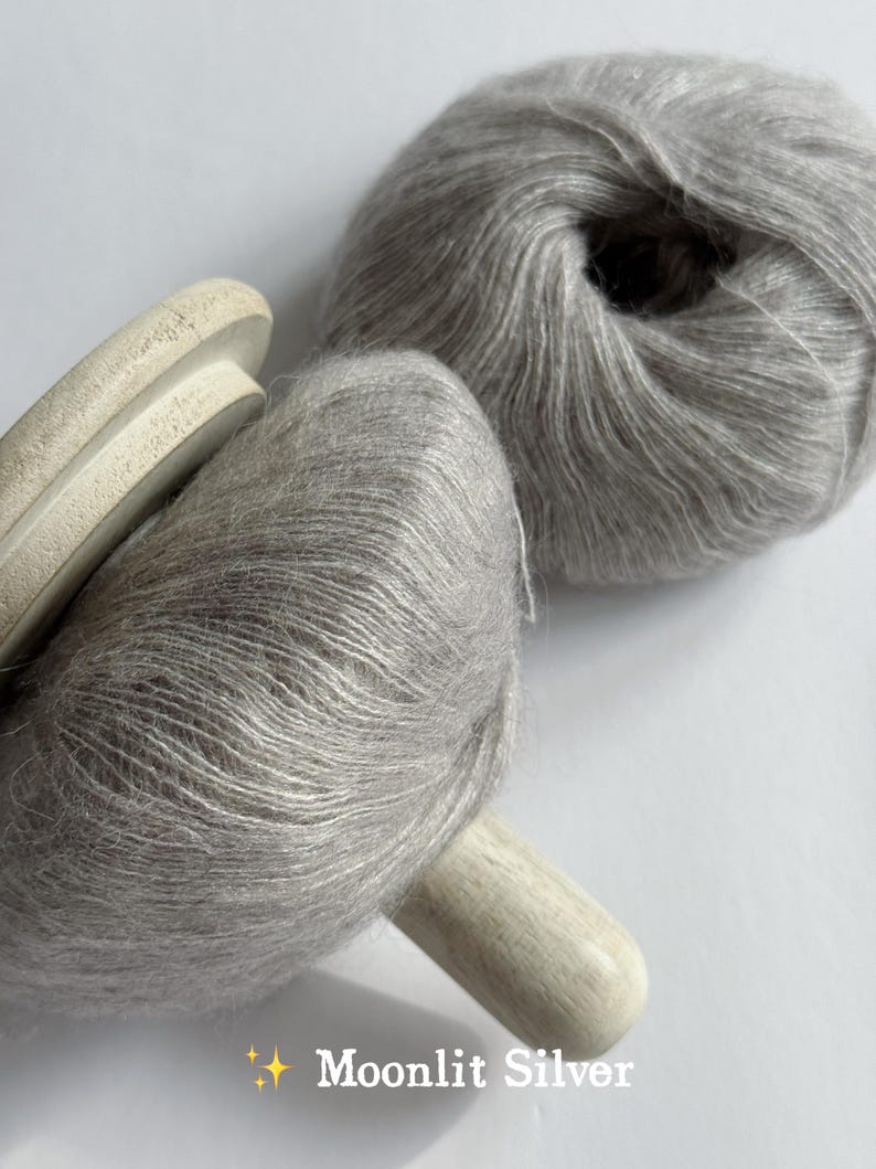 Puede incluir: Dos madejas de hilo Moonlit Silver, una en un carrete de madera. El hilo es de un color gris claro y suave, con una textura esponjosa. El texto "Moonlit Silver" est&aacute; en la parte inferior de la imagen.