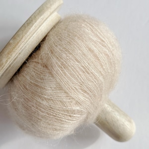 Puede incluir: Primer plano de una bola de hilo beige claro con una bobina de madera. El hilo tiene una textura suave y difusa. El texto "Champagne Glow" est&aacute; en la parte inferior de la imagen.