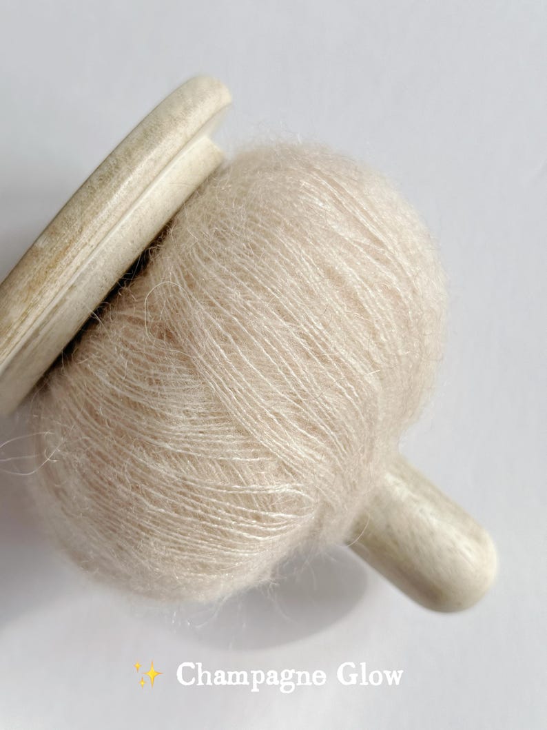 Puede incluir: Primer plano de una bola de hilo beige claro con una bobina de madera. El hilo tiene una textura suave y difusa. El texto "Champagne Glow" est&aacute; en la parte inferior de la imagen.