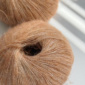 Puede incluir: Dos ovillos de hilo marr&oacute;n claro con una textura suave y esponjosa. El hilo tiene un brillo sutil y una apariencia delicada, perfecto para proyectos de tejido o ganchillo. El hilo es de un color c&aacute;lido y neutro.
