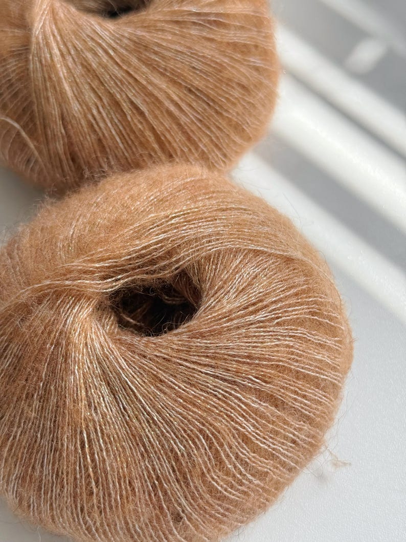 Puede incluir: Dos ovillos de hilo marr&oacute;n claro con una textura suave y esponjosa. El hilo tiene un brillo sutil y una apariencia delicada, perfecto para proyectos de tejido o ganchillo. El hilo es de un color c&aacute;lido y neutro.