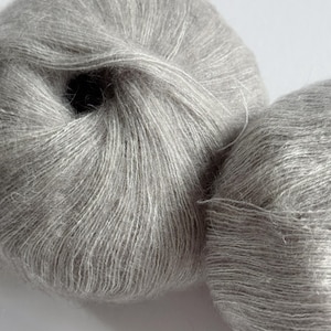 Puede incluir: Dos ovillos de hilo Moonlit Silver. El hilo es de un color gris claro y suave. El texto "Moonlit Silver" se muestra en blanco en la parte inferior de la imagen.