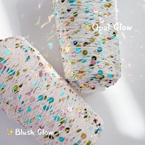 May include: Two spools of white yarn, embellished with colourful sequins. One is labelled "Opal Glow" and the other "Blush Glow". The sequins are in shades of blue, pink, gold, and green, adding sparkle to the yarn.