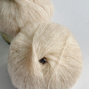 Puede incluir: Dos madejas de hilo Champagne Glow, de color beige claro y suave. El hilo est&aacute; enrollado en bolas redondas, con un husillo de madera visible en el fondo. El texto "Champagne Glow" est&aacute; en la parte inferior.
