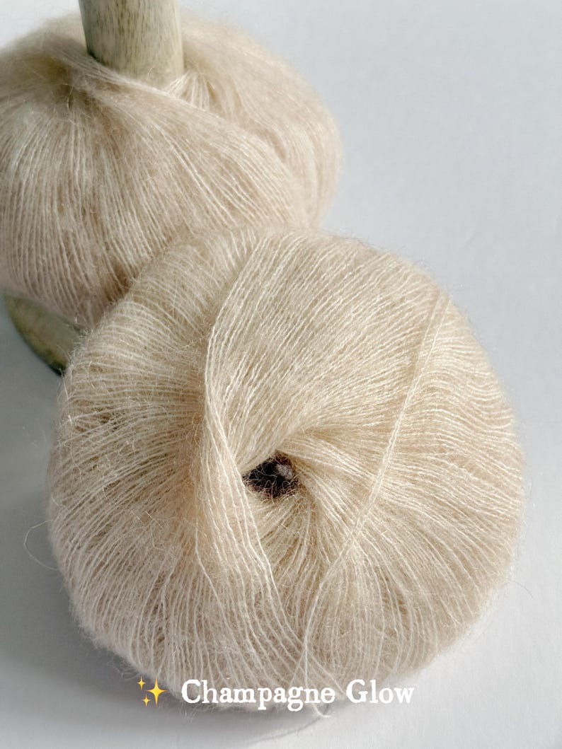 Puede incluir: Dos madejas de hilo Champagne Glow, de color beige claro y suave. El hilo est&aacute; enrollado en bolas redondas, con un husillo de madera visible en el fondo. El texto "Champagne Glow" est&aacute; en la parte inferior.