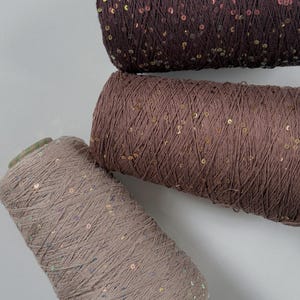 May include: Three spools of yarn in varying shades of brown. The yarn is embellished with small, iridescent sequins. The spools are arranged diagonally, showcasing the texture and color variations of the yarn.
