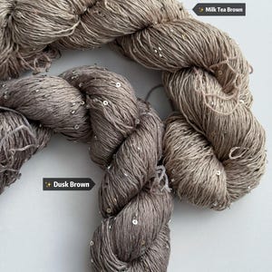 May include: Close-up of two skeins of yarn in shades of brown, with the labels "Milk Tea Brown" and "Dusk Brown." The yarn is embellished with small, shiny sequins. The yarn is tightly twisted, showing the texture and color variations.