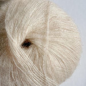 Puede incluir: Primer plano de un ovillo de hilo Champagne Glow. El hilo es de color beige claro y suave, con una textura delicada y difusa. El texto "Champagne Glow" est&aacute; impreso en una fuente cursiva en la parte inferior. Ideal para tejer y hacer ganchillo.