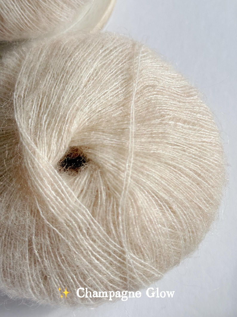 Puede incluir: Primer plano de un ovillo de hilo Champagne Glow. El hilo es de color beige claro y suave, con una textura delicada y difusa. El texto "Champagne Glow" est&aacute; impreso en una fuente cursiva en la parte inferior. Ideal para tejer y hacer ganchillo.