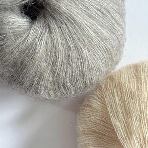 Puede incluir: Primer plano de dos ovillos de lana. Uno es de color gris claro y el otro es de color blanco roto cremoso. El hilo parece suave y esponjoso, con hebras visibles. Ideal para proyectos de punto o ganchillo.