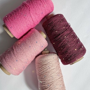 May include: Four spools of yarn in shades of pink and burgundy, each with a cardboard core. The yarn is tightly wound and has a textured appearance, with small, shiny sequins scattered throughout. The colors range from light pink to deep burgundy.