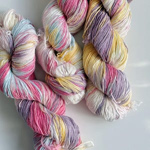 Puede incluir: Primer plano de varios ovillos de hilo colorido. El hilo es una mezcla de rosa, amarillo, morado y blanco. El hilo est&aacute; torcido y parece suave, ideal para proyectos de tejido o ganchillo.